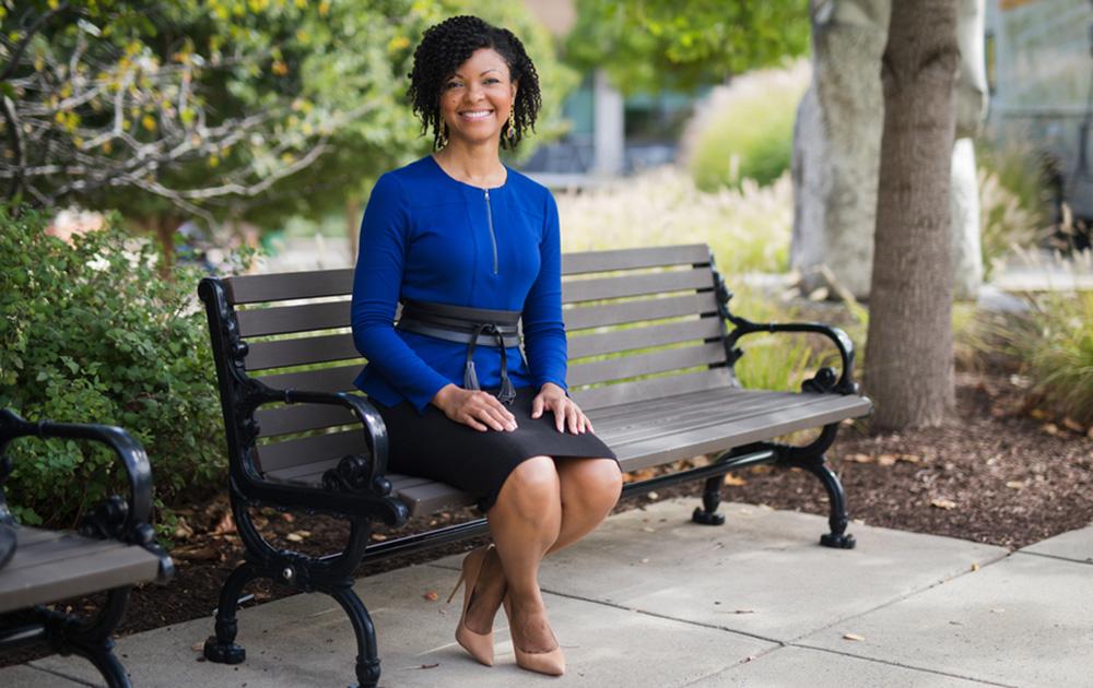 Terry-Ann Jones poses for a photo sitting on a bench. She wears a blue dress and smiles at the camera.